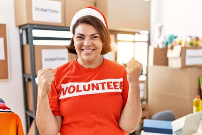 Middle age hispanic woman wearing volunteer t shirt and christmas hat screaming proud, celebrating victory and success very excited with raised arms